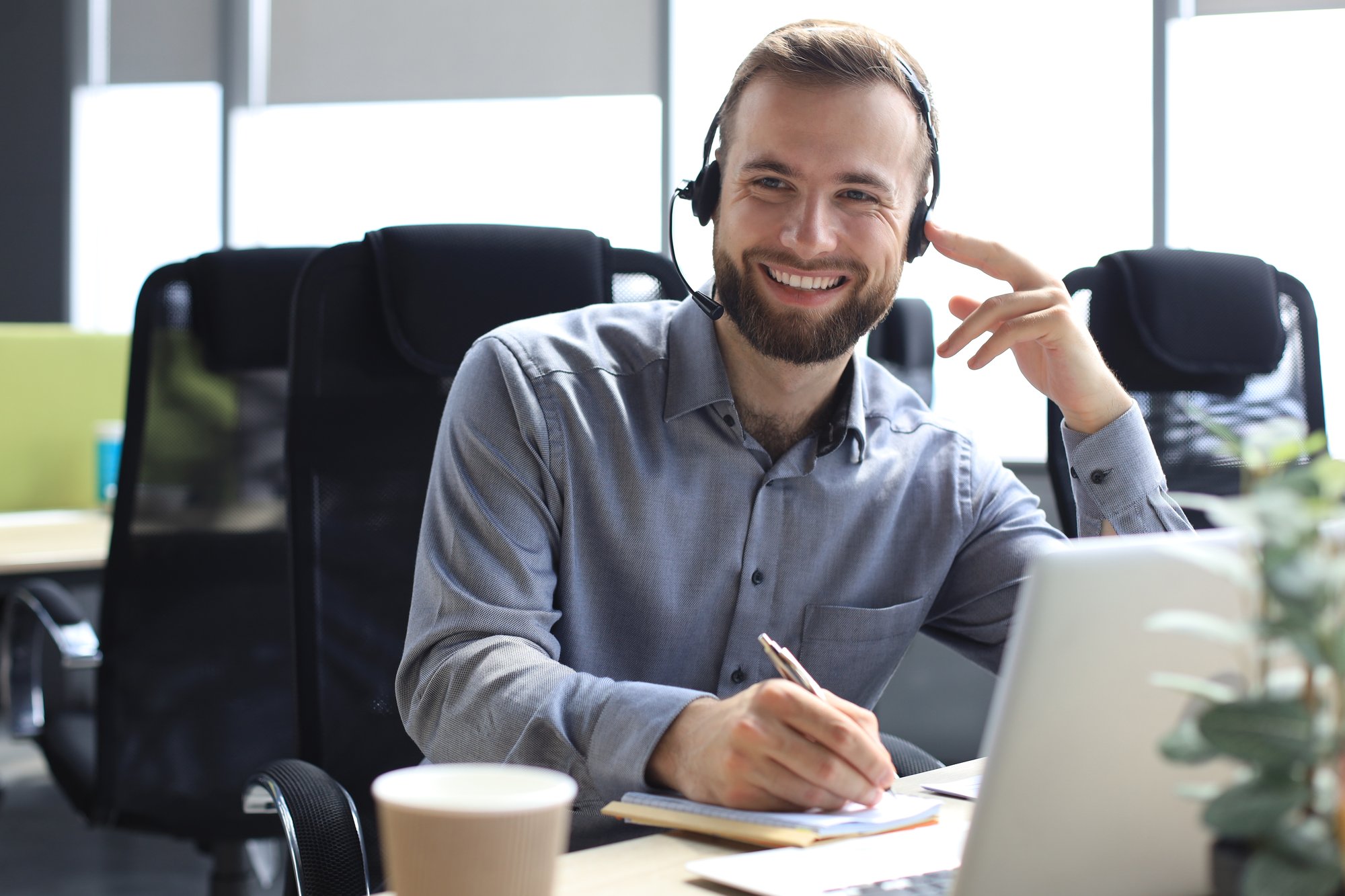operador-de-call-center-masculino-sorridente-com-fones-de-ouvido-sentado-em-um-escritorio-moderno-consultando-informacoes-line-em-um-laptop-procurando-informacoes-em-um-arquivo-para-ajudar-o-cliente
